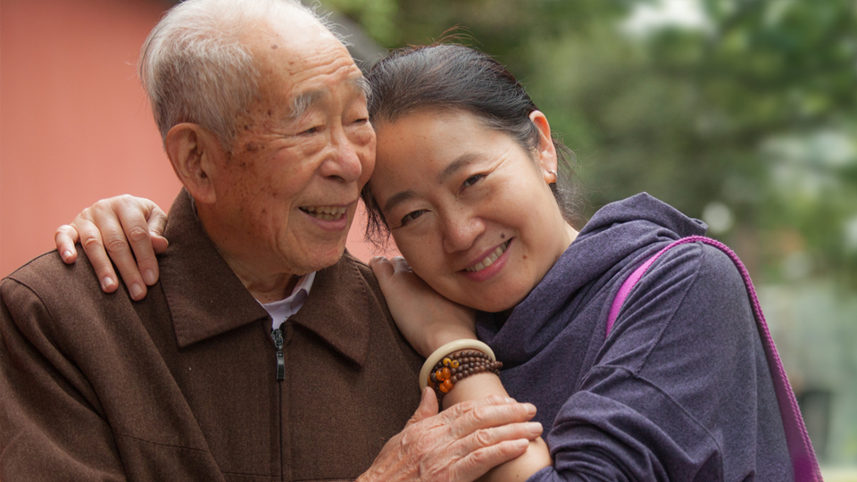 Cancer System Quality Index 2020 Adult woman leaning in to her elderly father's shoulder, with an arm around him, smiling at the camera.